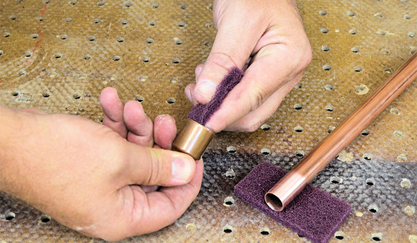 A worker uses a piece of a non-woven abrasive pad to clean a copper pipe