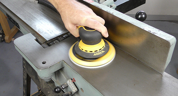 A worker sands the surface of a planer table with a non-abrasive pad mounted to a random orbit sander