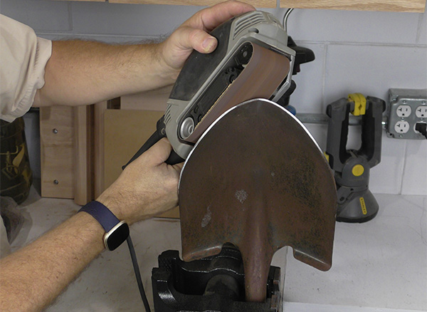 A worker uses a belt sander to clean and sharpen the edges of a spade.