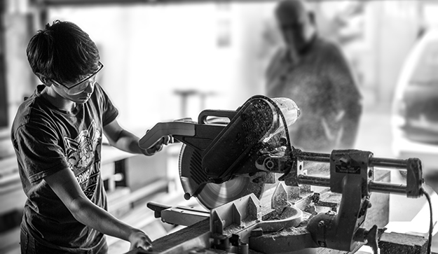 A boy works on power tools in a wood shop while his father  watches. Photo by Vance Osterhout on Unsplash